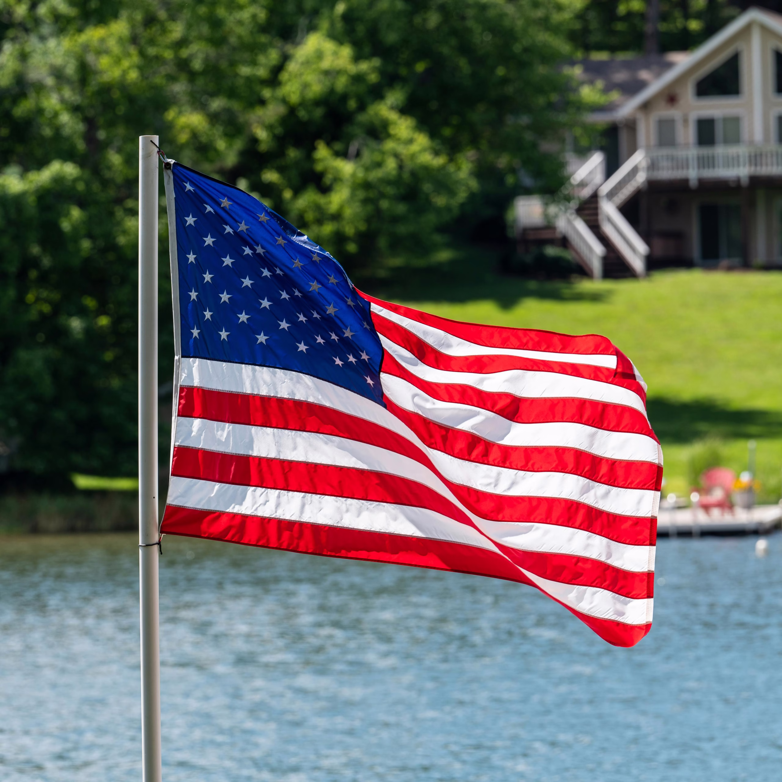Vibrant American flag waving by a picturesque lake with a house backdrop, capturing summer spirit.